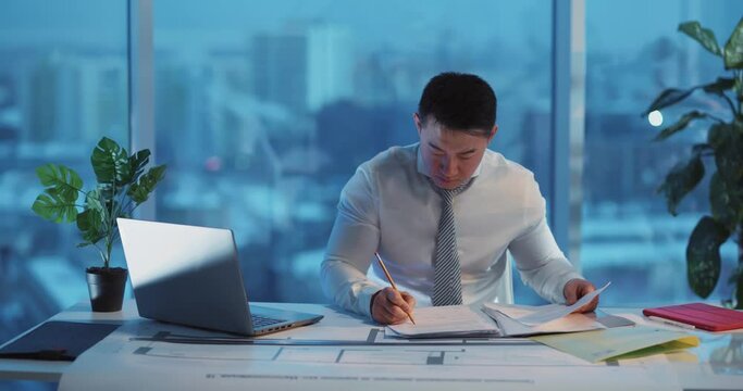 Asian young businessman sitting in corporate cozy office department and doing buisness tasks solving financial issues performing paperwork.