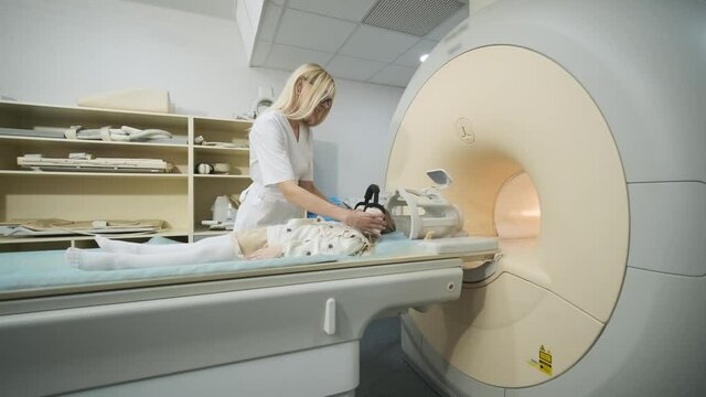 Doctor radiologist puts noise isolating headphones on the patient little girl to make an MRI scanning of brain, head, neck, girl lies on automatic table, using modern equipment, coil on the head