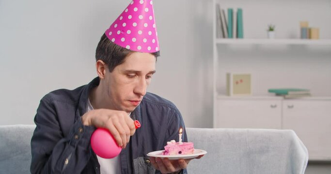 Portrnt single millennial man celebrating his birthday alone at home during quarantine wears pink hat on his head blowing up birthday candle on cake using balloon pretending happy feeling desperate
