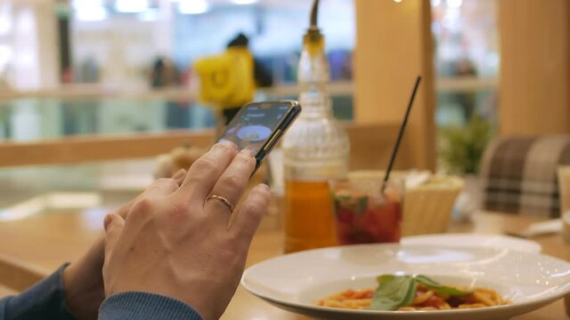 In the cafe, a visitor sits at a wooden table, Italian pasta and a fruit cocktail on the table. A visitor drives a smartphone screen to photograph food. Close-up.