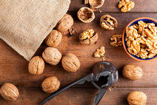 Flat Lay Image Showing A Cast Iron Walnut Pliers ( A Nutcracker Tool) With Raw Fresh Walnuts In Shell On Wooden Table. A Small Bowl Of Walnut Kernels And Some Shells Are Seen As Well As A Burlap Sack.