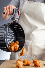 A woman is holding the handle of an air fryer oven basket with homemade fresh breaded chicken nuggets. She is pouring them onto baking paper over kitchen counter. A convenient low fat frying tool.