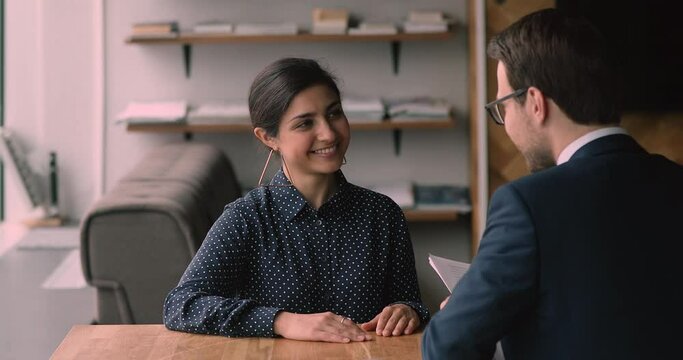 Smiling indian businesswoman holding negotiations meeting, sitting at table opposite male business partner. Skilled young mixed race woman passing job interview with confident leader employer.