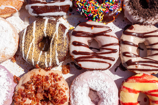 Closeup angled image of fresh made store bought donuts in paper box fresh out of the donut shop. An assortment with different flavors and toppings are packed together on flat box, a sweet summer treat