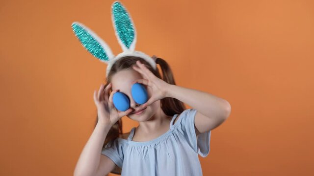 Happy Easter. A little Caucasian girl with bunny ears on her head closes her eyes with two blue eggs. Shakes his head in different directions.