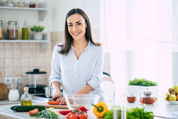 Cute happy young brunette woman in good mood preparing a fresh vegan salad for a healthy life in the kitchen of her home