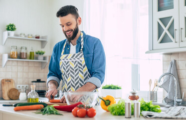 Fototapeta na wymiar Handsome smiling young bearded man cooks fresh healthy vegan salad for his family and having fun in the big light kitchen