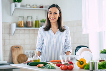 Cute happy young brunette woman in good mood preparing a fresh vegan salad for a healthy life in the kitchen of her home