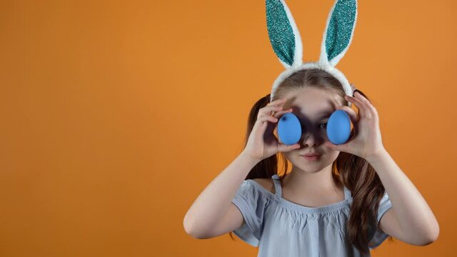 Happy Easter. A little Caucasian girl with rabbit ears on her head holds two blue eggs in her hands, closes and opens her eyes with them.