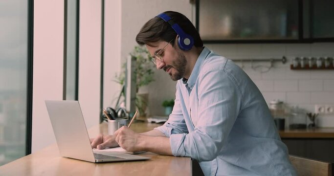 Focused young 30s businessman in eyewear and headphones looking at laptop screen, watching educational seminar or taking part in negotiations meeting, writing notes in copybook, e-learning concept.