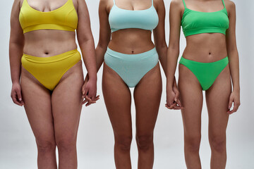 Cropped shot of three women with different body shapes in colorful underwear holding hands, supporting each other, standing together isolated over light background - obrazy, fototapety, plakaty