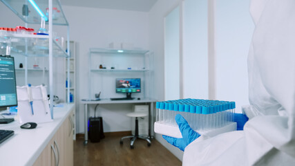 Chemist in protection suit bringing test tubes in steril laboratory and typing data on computer. Doctors examining virus evolution using high tech for research of treatment development against covid19