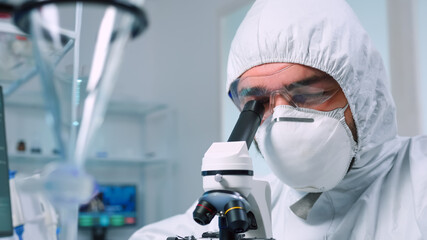 Biochemist in ppe suit looking through a microscope during experiment in sterile lab. Chemist researcher examining virus evolution using high tech for vaccine scientific research against covid19