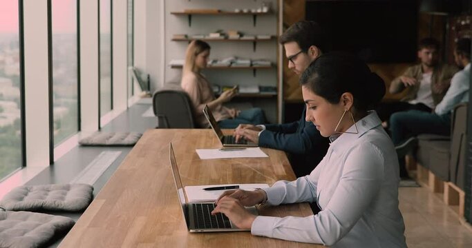 Focused young indian ethnicity businesswoman working on computer online, sitting at shared table in modern coworking open space place with diverse freelance people, office lifestyle concept.