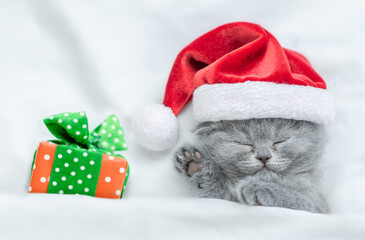 Cute kitten wearing red santa's hat sleeps with gift box under a white blanket on a bed. Top down...