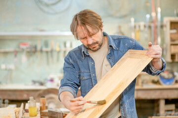Worker applies varnish on wooden detail with paintbrush in  carpentry workshop