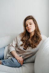 Obraz premium A young blonde woman in casual comfortable clothes sits on a white sofa at home. scandinavian interior, minimalism