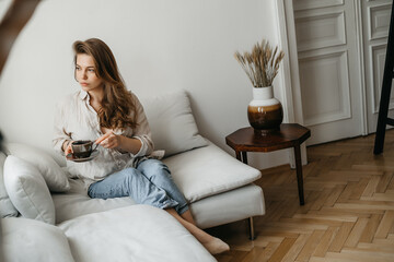 A young blonde woman in casual comfortable clothes sits on a white sofa at home and drinks coffee. scandinavian interior, minimalism