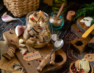 homemade marinated mushrooms in glass jar on wooden board with herbs, dill. pepper, vintage spoons