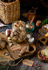 homemade marinated mushrooms in glass jar on wooden board with herbs, dill. pepper, vintage spoons