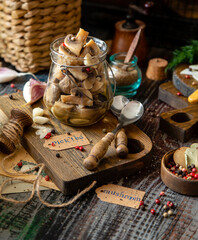 homemade marinated mushrooms in glass jar on wooden board with herbs, dill. pepper, vintage spoons