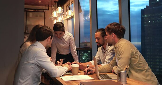Focused young creative mixed race international company employees discussing new corporate software mobile application with indian leader at brainstorming meeting in modern office with night city view