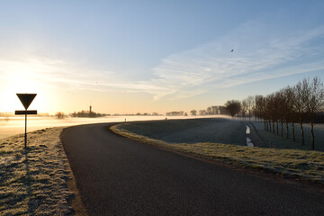 A rising morning sun shining on a winding Dutch road with a ground frost covering close to Oss,...