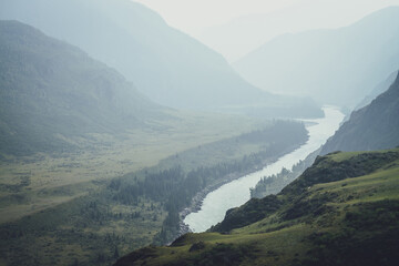 Misty mountain landscape with wide mountain river. Dark green gloomy scenery with confluence of two...
