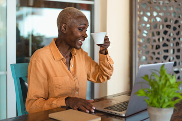 Afro senior woman having fun doing video call using computer while drinking coffee in bar restaurant