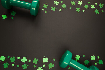Two heavy green dumbbells and shamrock clover for St. Patrick's Day. Healthy fitness gym flat lay composition concept, with copyspace on black background.