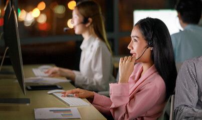 Portrait adult pretty asian woman who is helpdesk team wearing headphone looking at screen monitor and smiling with typing on keyboard in night bokeh on blur background