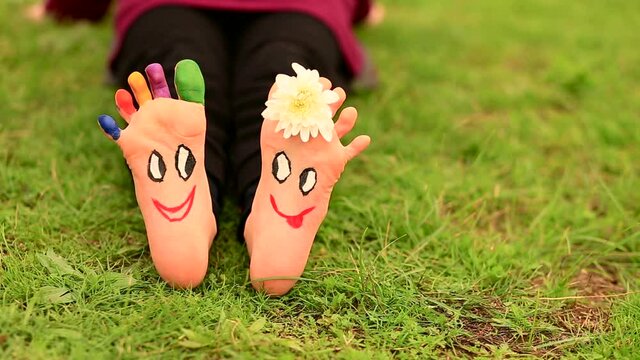 Little child girl sits on green grass with painted feet with funny comic faces 