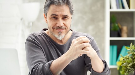 Portrait of man at home sitting at desk, working, looking at camera. Happy smile, grey hair, beard. Portrait of mature age, middle age, mid adult man in 50s.