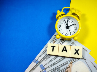 Selective focus.Clock and dollar banknotes with word TAX on blue and yellow background.Business concept.