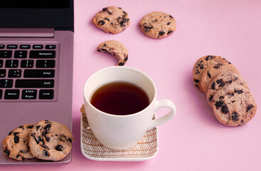 a cup of tea is on a saucer near a laptop. Cookies with chocolate on a pink background. Lifestyle concept in pink, glamorous workplace. High quality photo