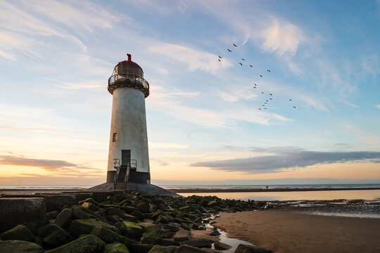 Beautiful lighthouse by sea golden hour sunset with rocks leading to stairs as ocean tide comes in.  Birds flying out to horizon sun reflecting in water pools of old abandoned amazing coastline beach