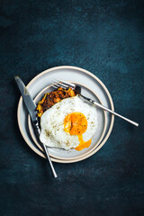 Vegetable zucchini and carrot fritters and an egg with a runny yolk on top, served on a blue plate, knife and fork, dark backdrop, top view