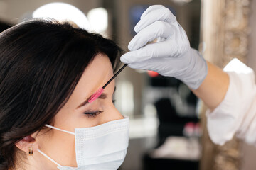 The professional eyebrow master is making an eyebrow correction for the girl in a medical mask in beauty salon. A beauty salon services during a pandemic.