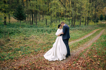 A beautiful bride with black hair and the groom are walking in the autumn park, hugging, enjoying each other.