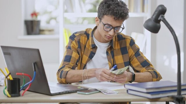 Confident concentrated Middle Eastern teenager in eyeglasses counting dollars and writing down income. Portrait of focused teenage boy controlling finances. Freelance and wealth. Slow motion.