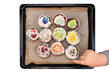 A child squeezes colored frosting from a tube onto chocolate brown cupcakes covered with white frosting with colorful decorations, isolated on white, top view.
