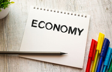 A notebook on a spring with the text ECONOMY lies on a wooden table with colored pens.