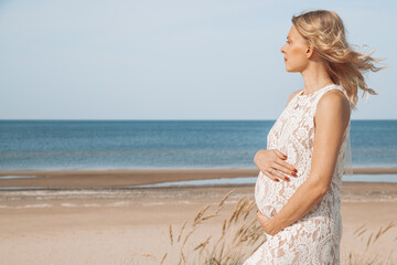 Fototapeta na wymiar Young, blond pregnant woman on the beach wearing white lace dress holding pregnancy belly. Free space. Maternity.