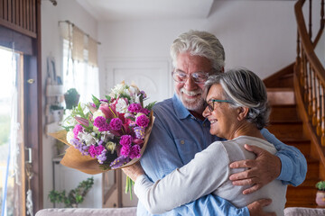 Portrait of couple of two happy and in love seniors or mature and old people holding flowers at home looking outside. Pensioners adult enjoying and celebrating holiday together. - Powered by Adobe