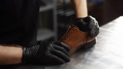 Close-up of shoemaker craftsman wearing black gloves inserts wooden shoe block into worn light brown leather shoes to be repaired in dark craft shoe shop. Concept of cobbler artisan work.