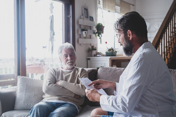 Male professional doctor consulting senior patient during medical care visit. Young man physician and old mature senior talking providing medical assistance sitting on sofa. Elderly people home care 
