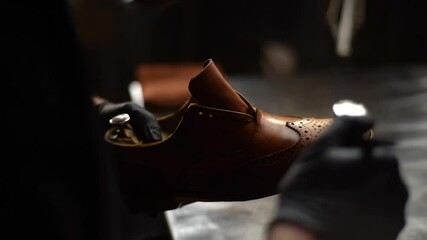 Hand held shot of shoemaker in black gloves spraying paint of light brown leather shoes, back view. Process of repairing and restoration shoes by cobbler in shoe repair shop with dark interior.