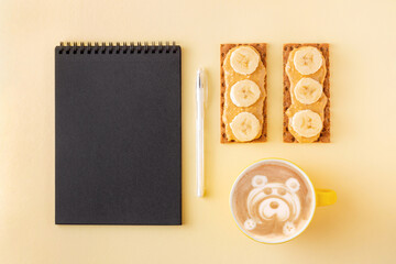 Planning during breakfast with cappuccino and crisp grain crackers with nut butter on yellow background. Wholegrain crispbread with nut paste and banana for morning coffee near notepad.