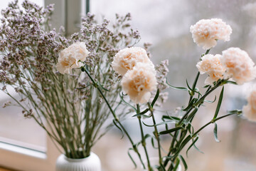 Beige carnations and dried flowers in a white vase on the windowsill