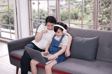 Smiling Asian mother and little girl child is relaxing with headphone and laptop for listening to online music or cartoon movie in social media on sofa in living room. Education and technology concept